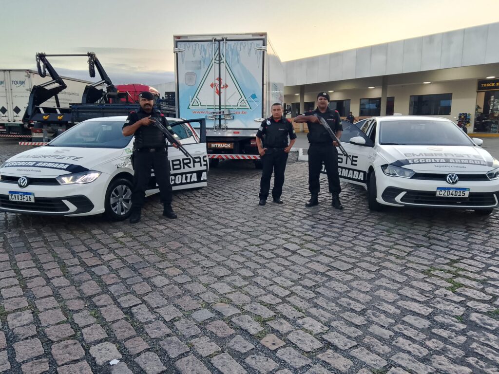 Três agentes de segurança armada da Embraste escoltam carga com dois veículos identificados, em pátio de parada rodoviária, ao lado de caminhões e estrutura de apoio logístico.
