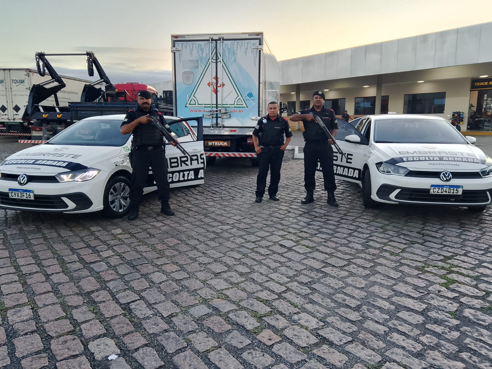 Três agentes de segurança armada da Embraste escoltam carga com dois veículos identificados, em pátio de parada rodoviária, ao lado de caminhões e estrutura de apoio logístico.