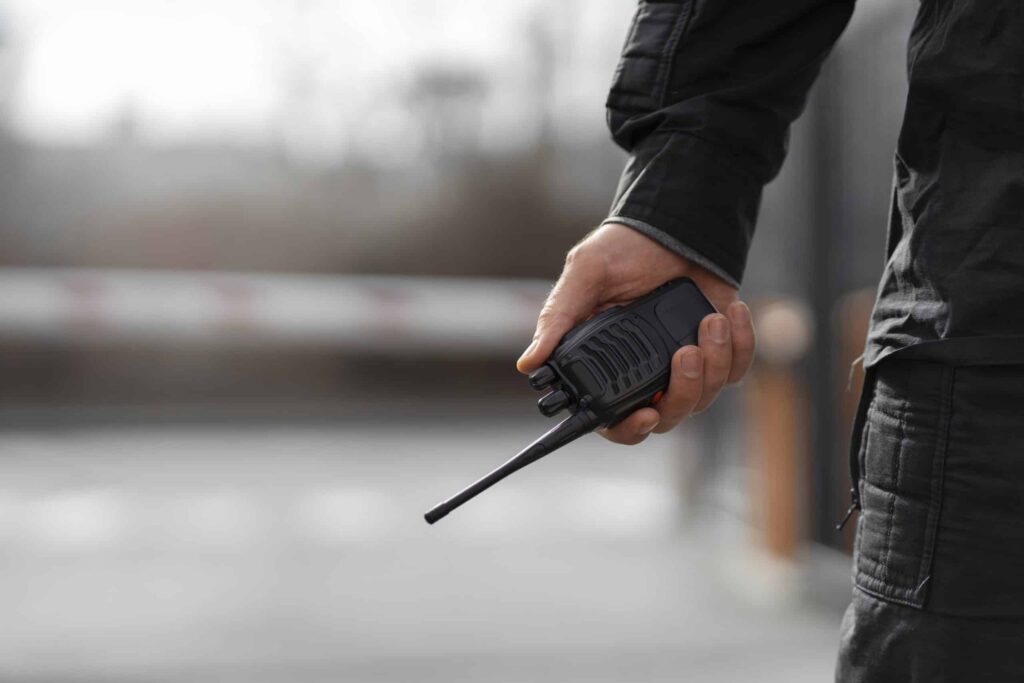 Segurança Patrimonial – Vigilante segurando rádio comunicador em área externa.