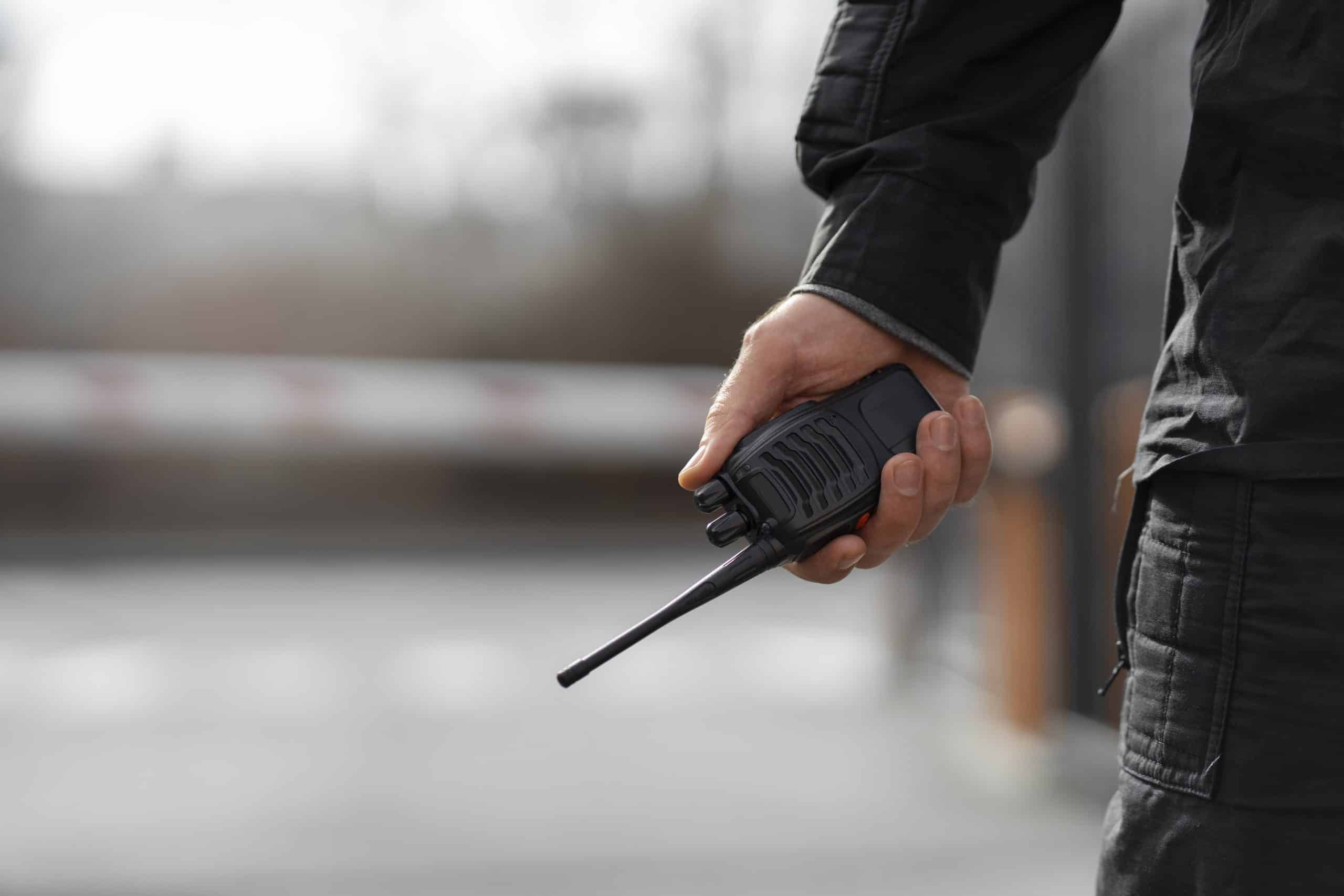 Segurança Patrimonial – Vigilante segurando rádio comunicador em área externa.
